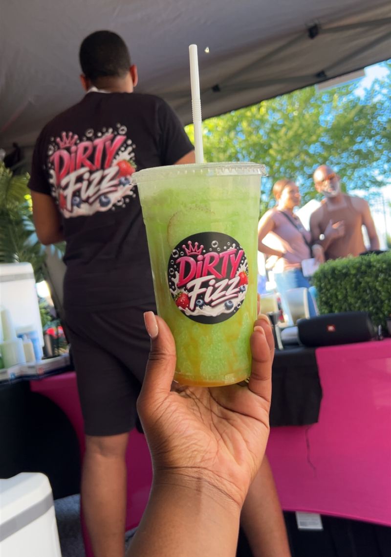 A hand holding a green Dirty Fizz drink in front of the bar